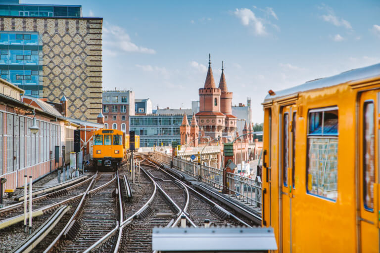 Blick auf zwei gelbe U-Bahn-Züge auf einer Brücke in Berlin, mit der markanten Architektur des Oberbaumbrücke im Hintergrund und modernen Gebäuden im Vordergrund.