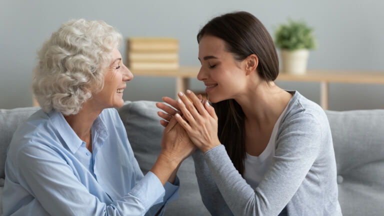 Junge Frau und ältere Dame lächeln sich an und halten sich sanft an den Händen, was eine herzliche Verbindung zwischen den Generationen symbolisiert. Der Hintergrund zeigt eine moderne Wohnatmosphäre mit Pflanzen und neutralen Farben.