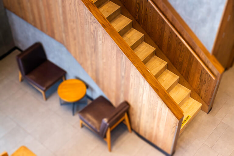 Holztreppe in modernem Innenraum mit zwei gemütlichen Sesseln und einem runden Tisch, ideal für entspannte Gespräche. Die Treppe ist gut beleuchtet und hat einen klaren Hinweis auf die Stufen.