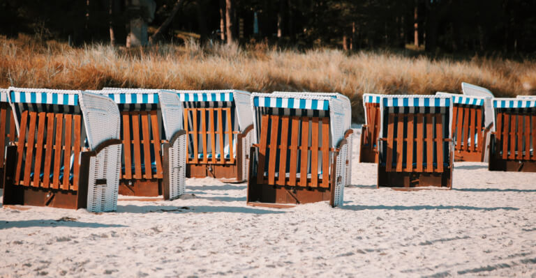 Strandkörbe in einer Reihe auf einem Sandstrand, umgeben von leichtem Gras und Bäumen im Hintergrund, ideal für entspannte Tage am Meer.