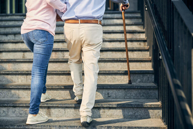 Eine Person mit Gehstock wird von einer Begleitperson beim Treppensteigen unterstützt, was die Bedeutung von Hilfe im Alltag und die Unterstützung älterer Menschen verdeutlicht.