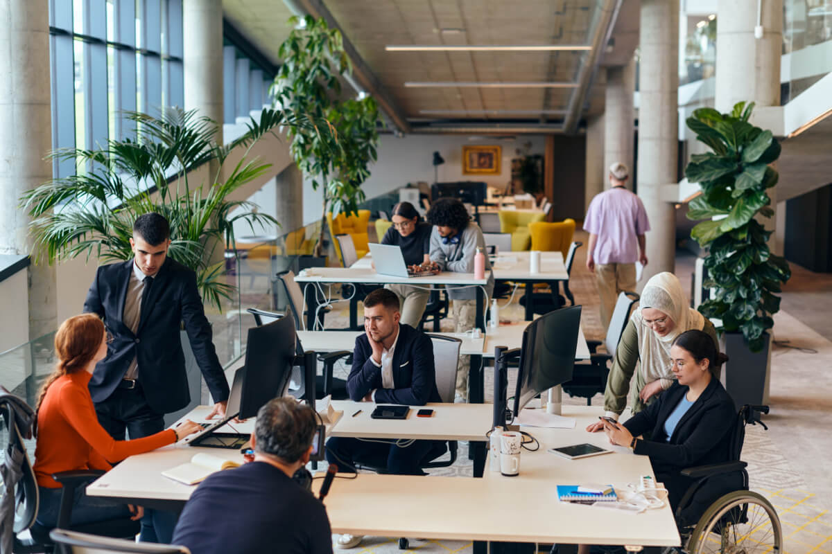 Moderne Büroumgebung mit mehreren Arbeitsplätzen, in der Mitarbeiter an Laptops und Monitoren arbeiten. Eine Frau in einem Rollstuhl wird von einer Kollegin unterstützt, während im Hintergrund weitere Personen an ihren Projekten arbeiten. Grünes Pflanzenarrangement sorgt für eine angenehme Atmosphäre.