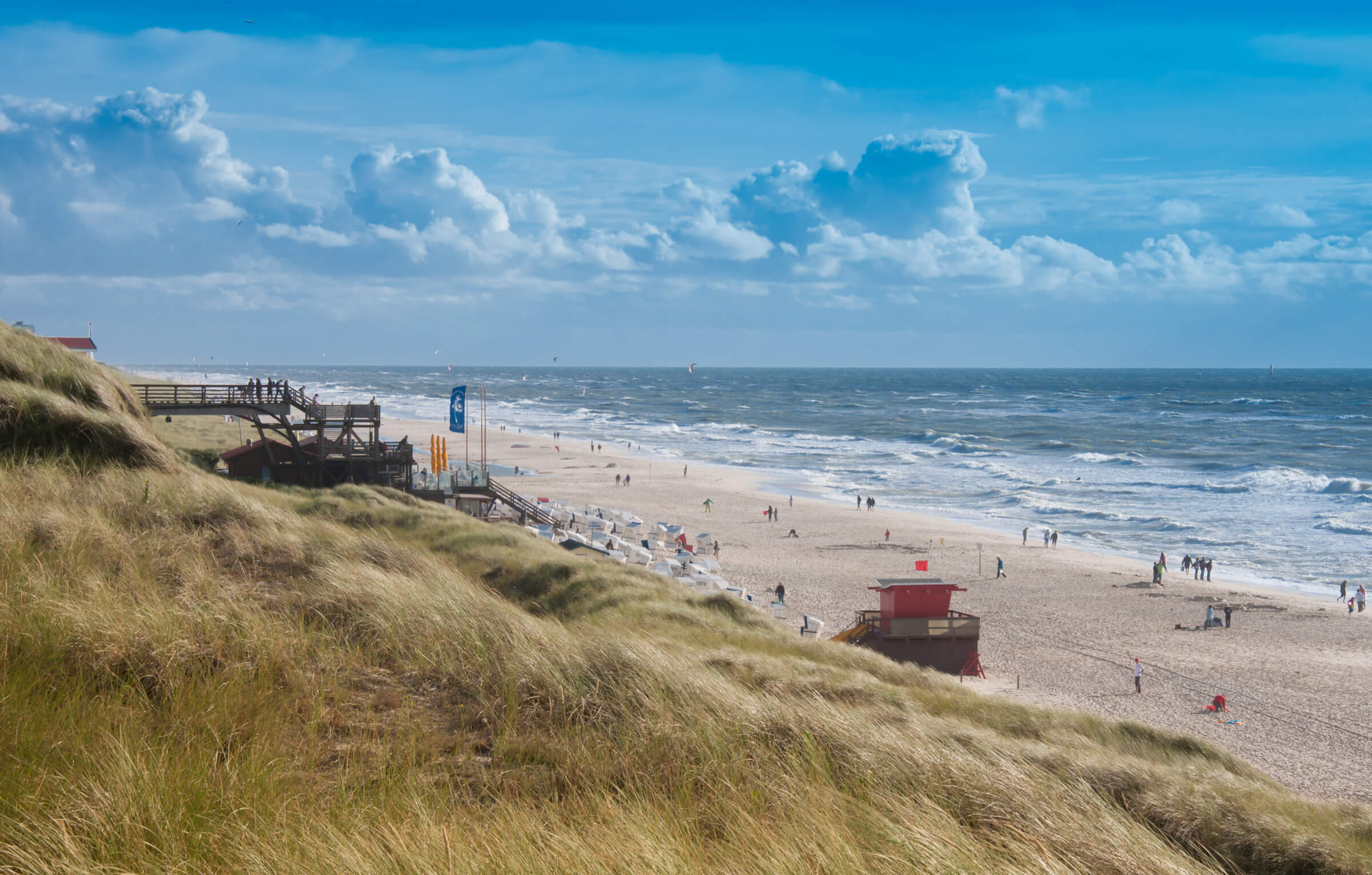 Blick auf einen weitläufigen Strand mit feinem Sand, sanften Wellen und einer lebhaften Küstenlandschaft, umgeben von Dünen und grasbewachsenen Hügeln. Im Hintergrund sind Menschen zu sehen, die am Strand spazieren gehen und sich entspannen, während farbige Flaggen und Strandkörbe die Szenerie ergänzen.
