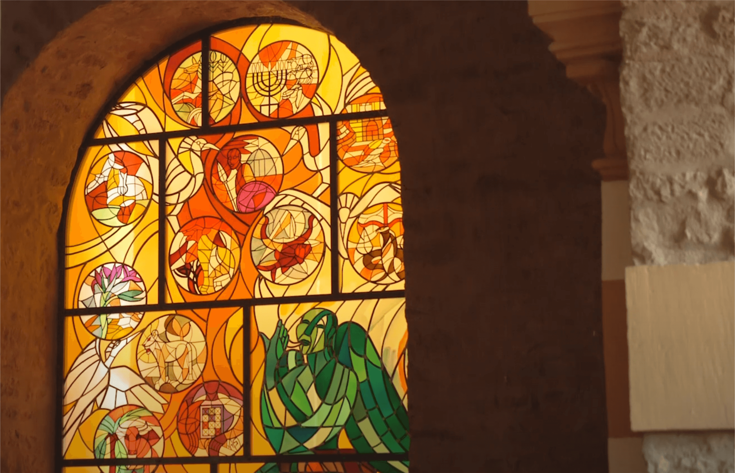 Buntes Kirchenfenster mit verschiedenen biblischen Motiven in warmen Orangetönen, das Licht durchlässt und die kunstvolle Gestaltung des Glases betont.
