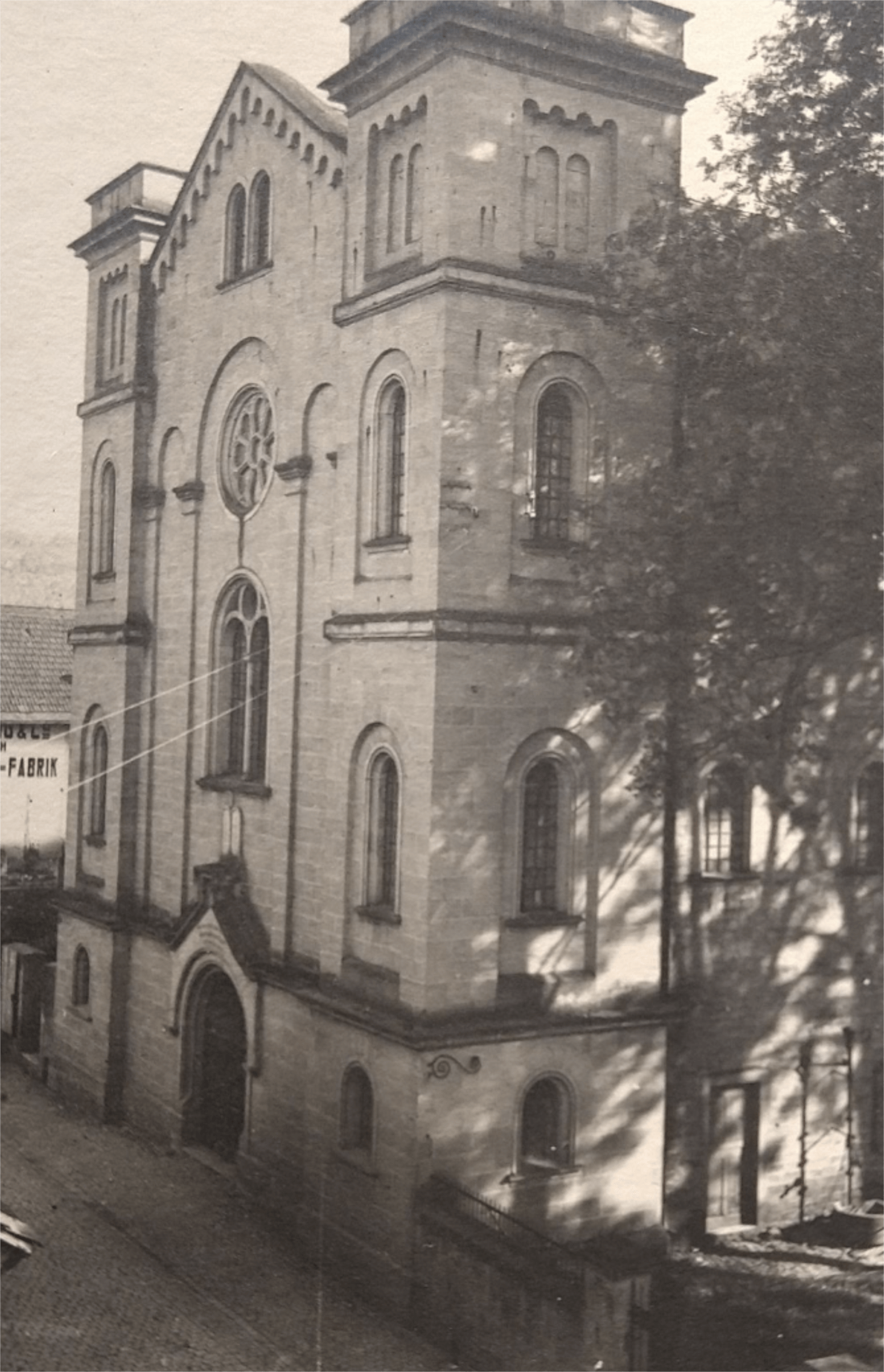 Historisches Gebäude einer ehemaligen Kirche, mit markanten Türmen und einem großen Rundfenster, umgeben von Bäumen und einer gepflasterten Straße. Im Hintergrund sind Teile eines alten Fabrikgebäudes sichtbar.