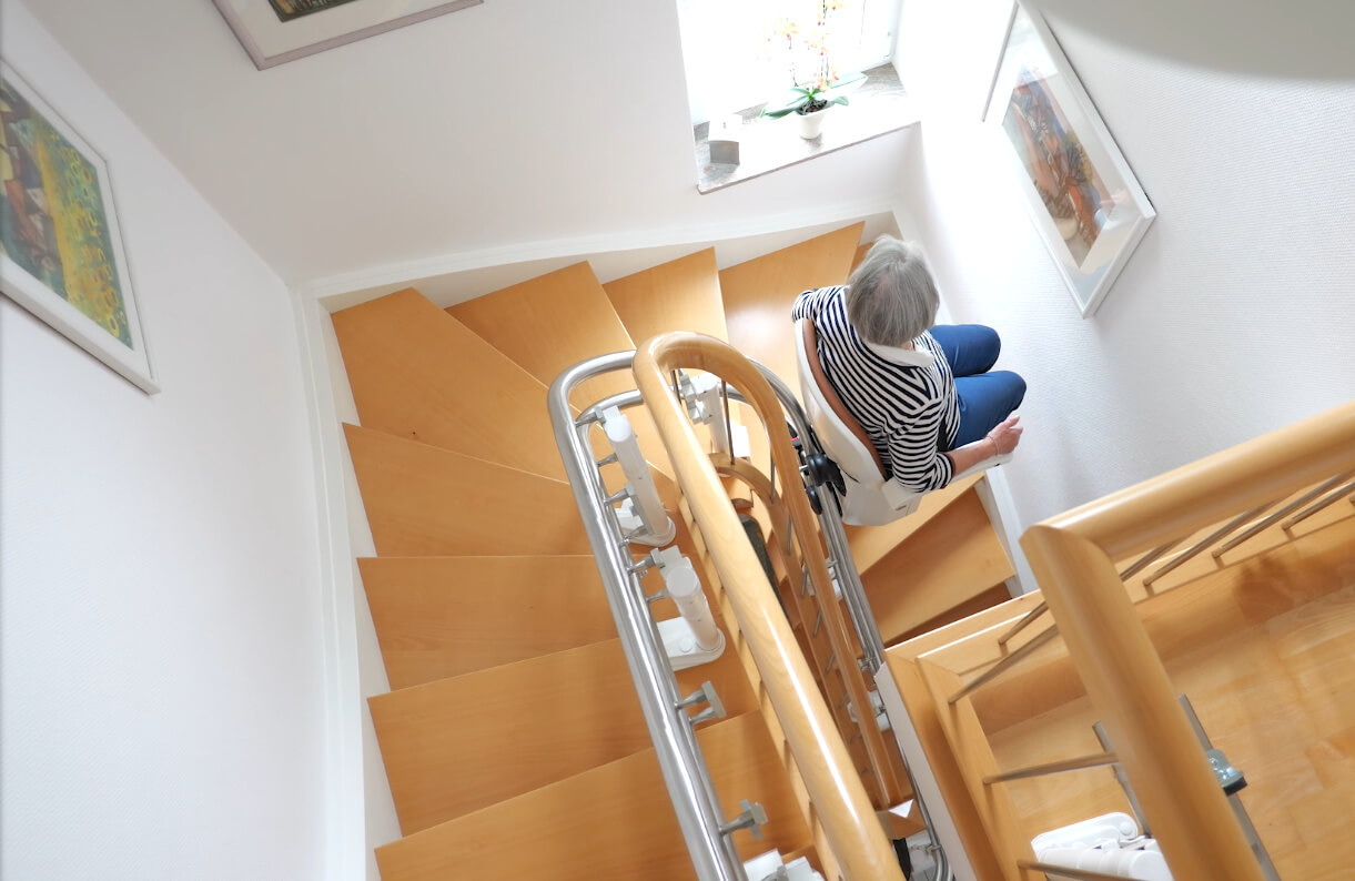 Eine ältere Person sitzt auf einem Treppenlift, der eine Wendeltreppe hinunterführt. Im Hintergrund sind helle Wände und Holztreppen zu sehen, während ein Fenster mit Pflanzen Licht in den Raum bringt.