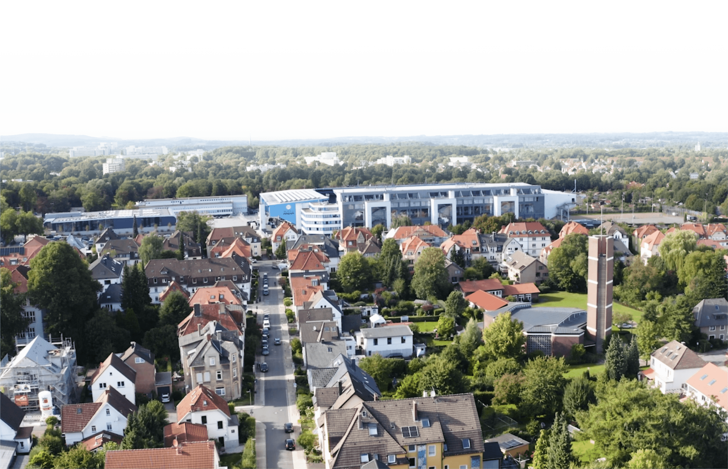 Luftaufnahme einer Wohngegend mit Einfamilienhäusern und mehrgeschossigen Gebäuden, umgeben von Bäumen und Grünflächen, im Hintergrund ein modernes Industriegebäude.