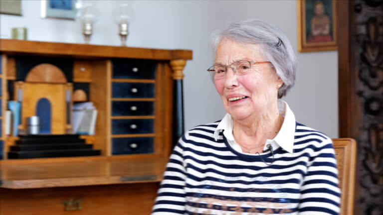 Portrait einer älteren Frau mit Brille, die in einem gemütlichen Raum lächelt. Im Hintergrund ist ein stilvoller Sekretär mit Holzoberfläche und verschiedenen Gegenständen zu sehen. Die Frau trägt ein gestreiftes Oberteil und hat graue Haare.