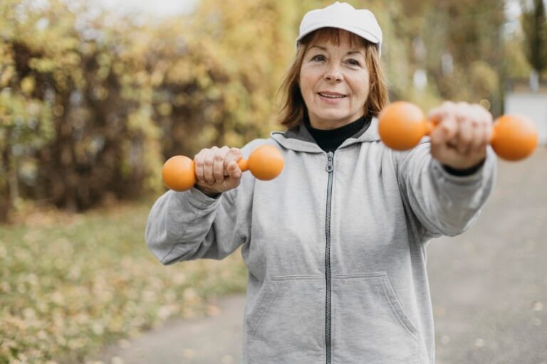Ältere Frau in sportlicher Kleidung mit einer grauen Jacke und einer weißen Kappe, die orangefarbene Hanteln hält und im Freien trainiert. Der Hintergrund zeigt herbstliche Bäume und einen Gehweg.