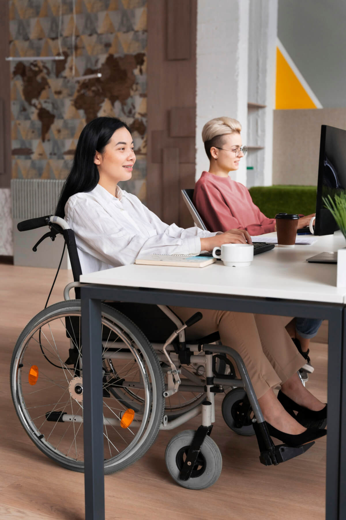 Zwei Frauen arbeiten in einem modernen Büro. Eine Frau sitzt im Rollstuhl und lächelt, während sie an einem Computer arbeitet. Die andere Frau sitzt an einem Schreibtisch und nutzt ebenfalls einen Computer. Im Hintergrund ist eine dekorative Wand mit einer Weltkarte zu sehen.